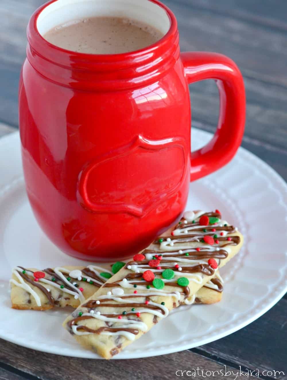 Cookie sticks drizzled with chocolate and white chocolate and topped with Christmas sprinkles beside a red mug of cocoa