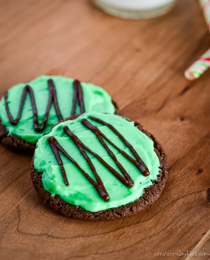 two chocolate cookies on a wood cutting board frosted with green mint frosting and drizzled with melted chocolate