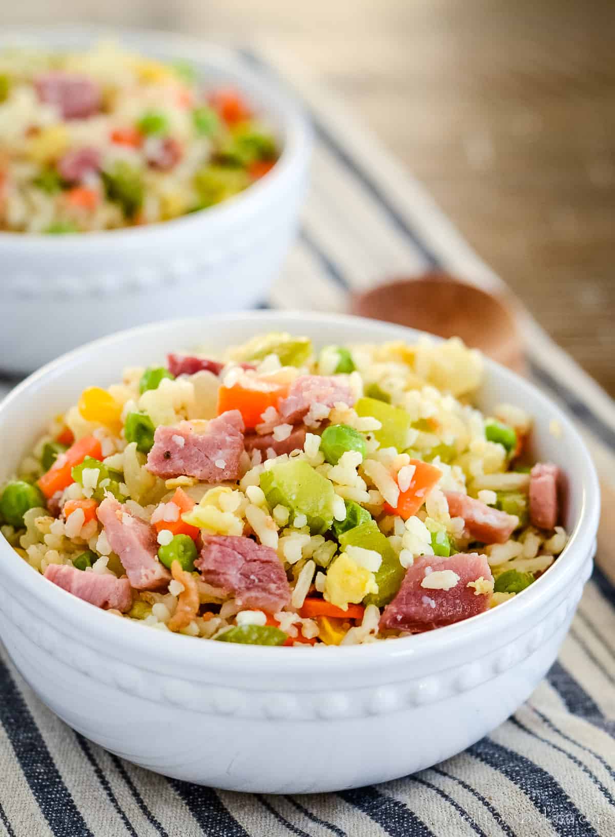 two bowls of ham fried rice on a striped dish towel with a wooden spoon