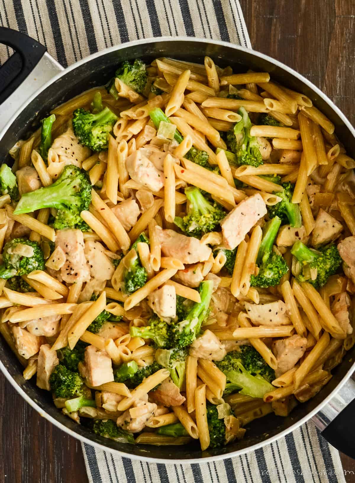 overhead shot of skillet full of chicken broccoli pasta on top of a striped dish towel