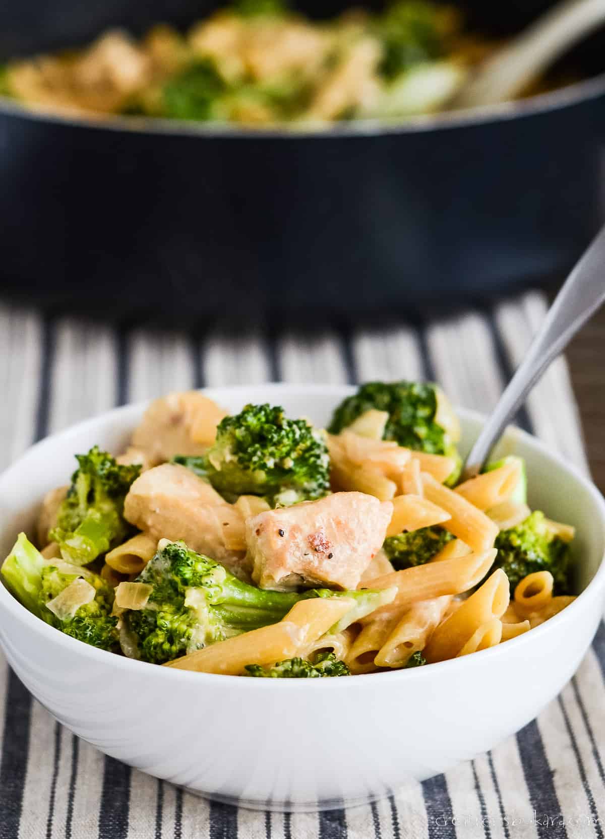 bowl of broccoli chicken pasta on a striped dish towel with a skillet in the background