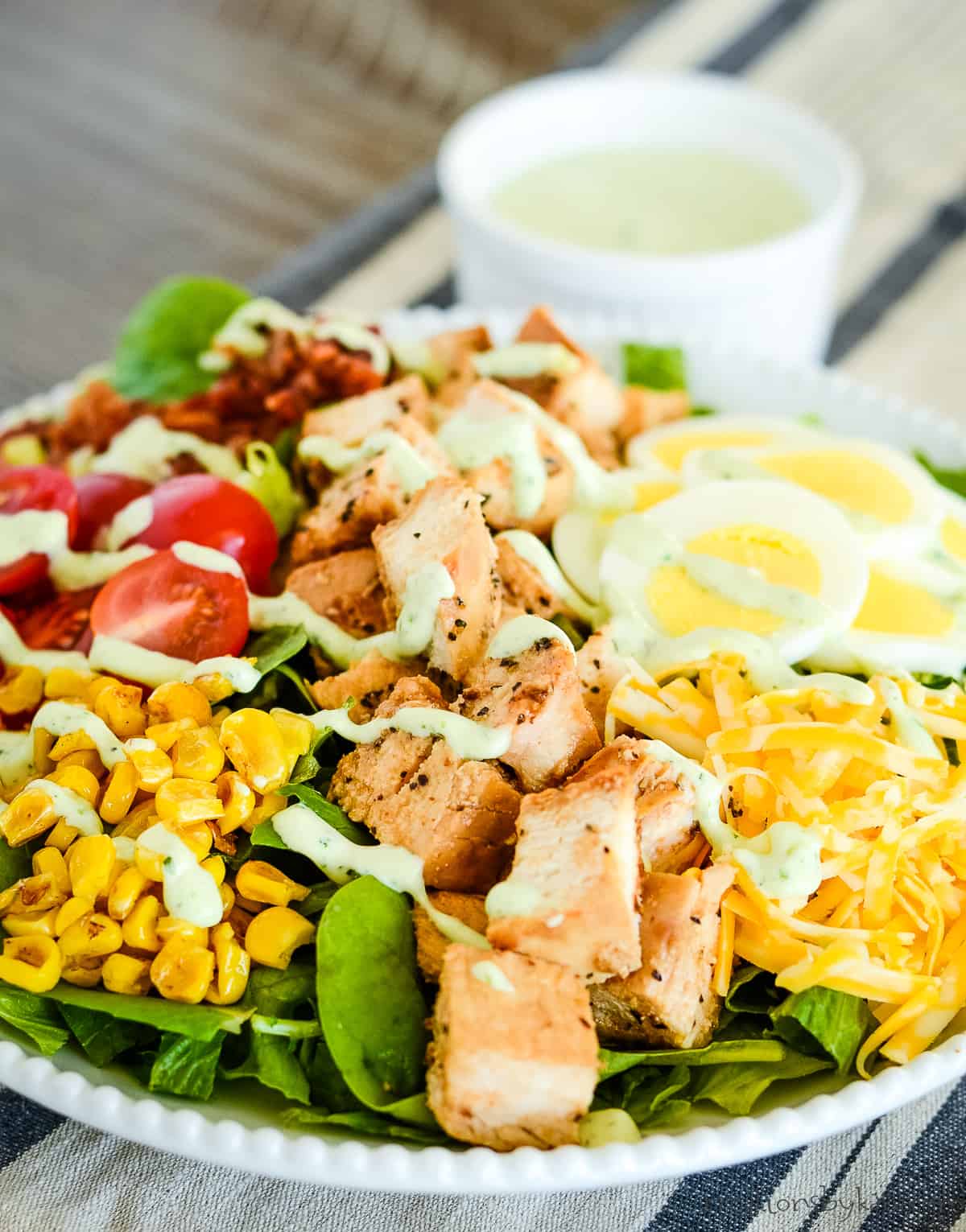 plate of salad with grilled chicken on a striped tablecloth with a bowl of dressing in the background