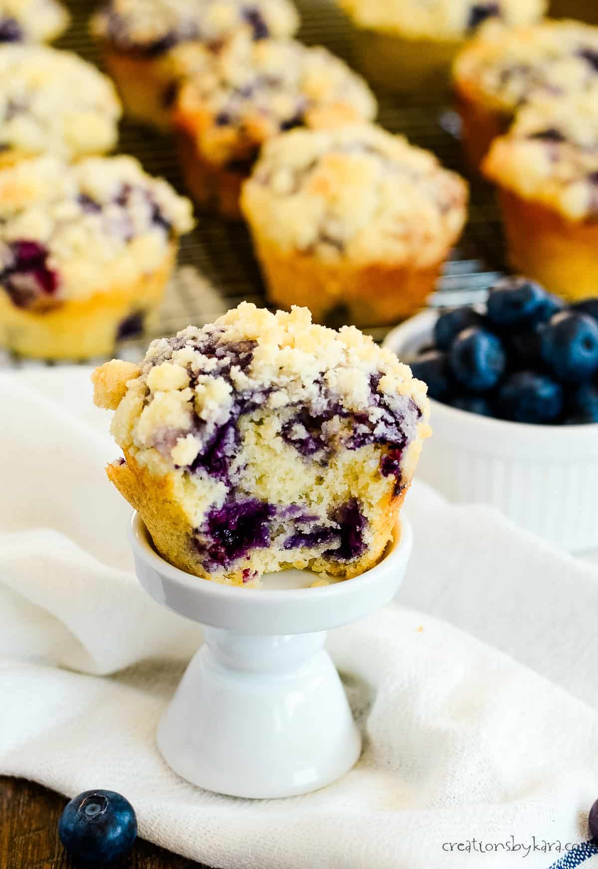 crumb topped blueberry muffin on a white stand with muffins and blueberries in the background
